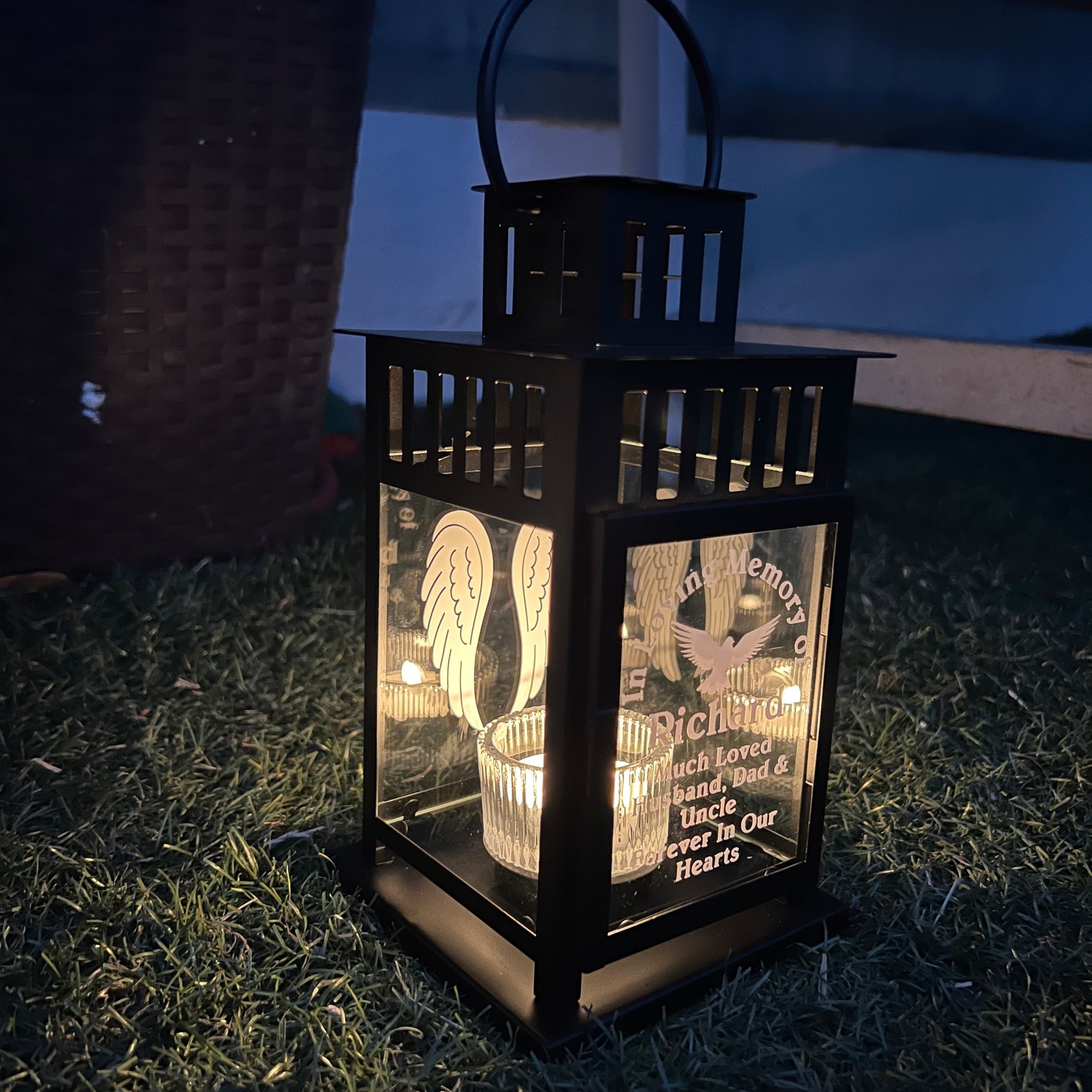 A black outdoor lantern with a glass window displaying a printed memorial-style design, placed on grass at night.