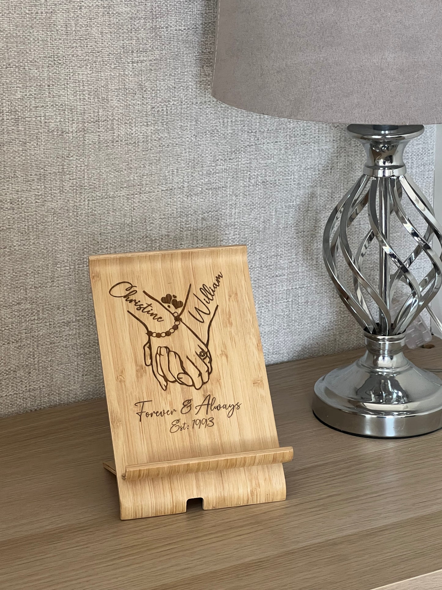 A unique personalised bamboo phone stand laser-engraved with a couple holding hands, two names & a date. Perfect wedding, anniversary or Valentine's gift.