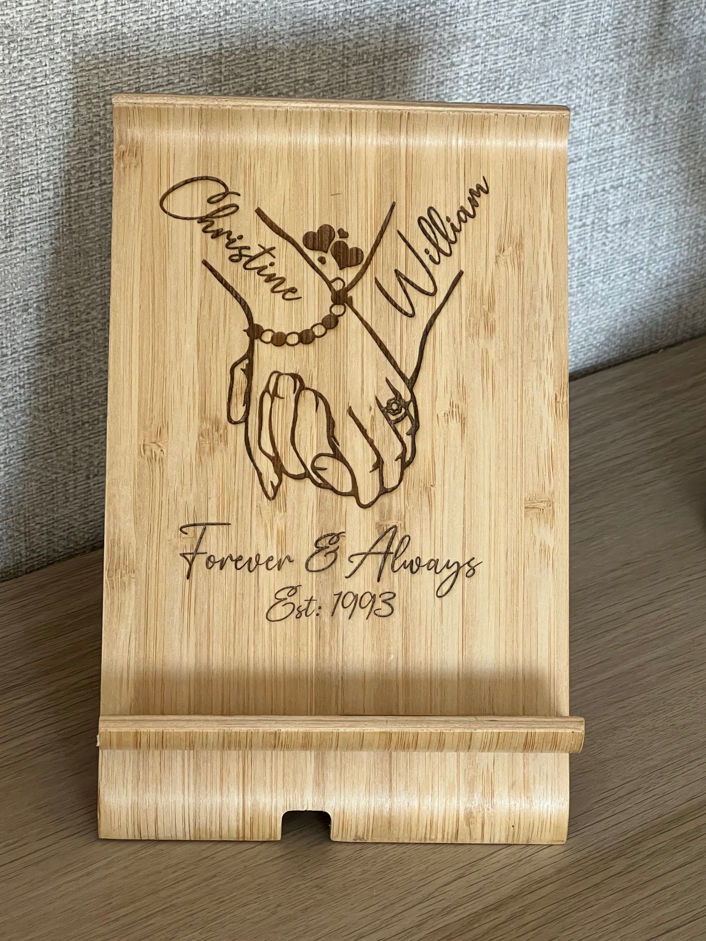 A unique personalised bamboo phone stand laser-engraved with a couple holding hands, two names & a date. Perfect wedding, anniversary or Valentine's gift.