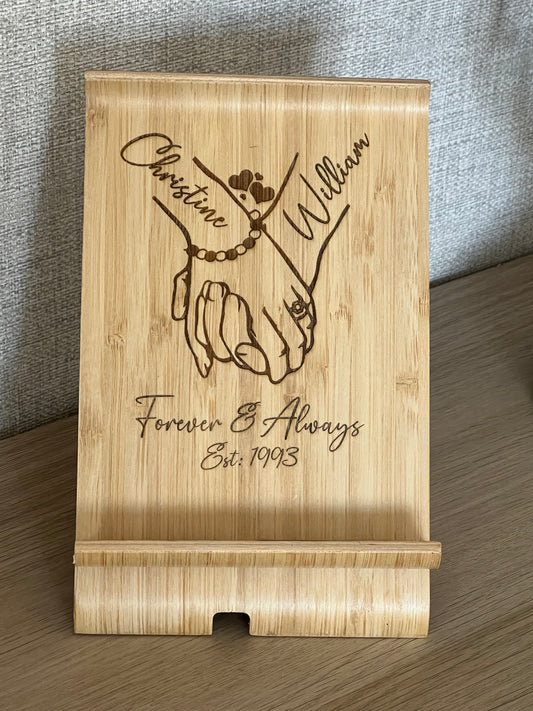 A unique personalised bamboo phone stand laser-engraved with a couple holding hands, two names & a date. Perfect wedding, anniversary or Valentine's gift.