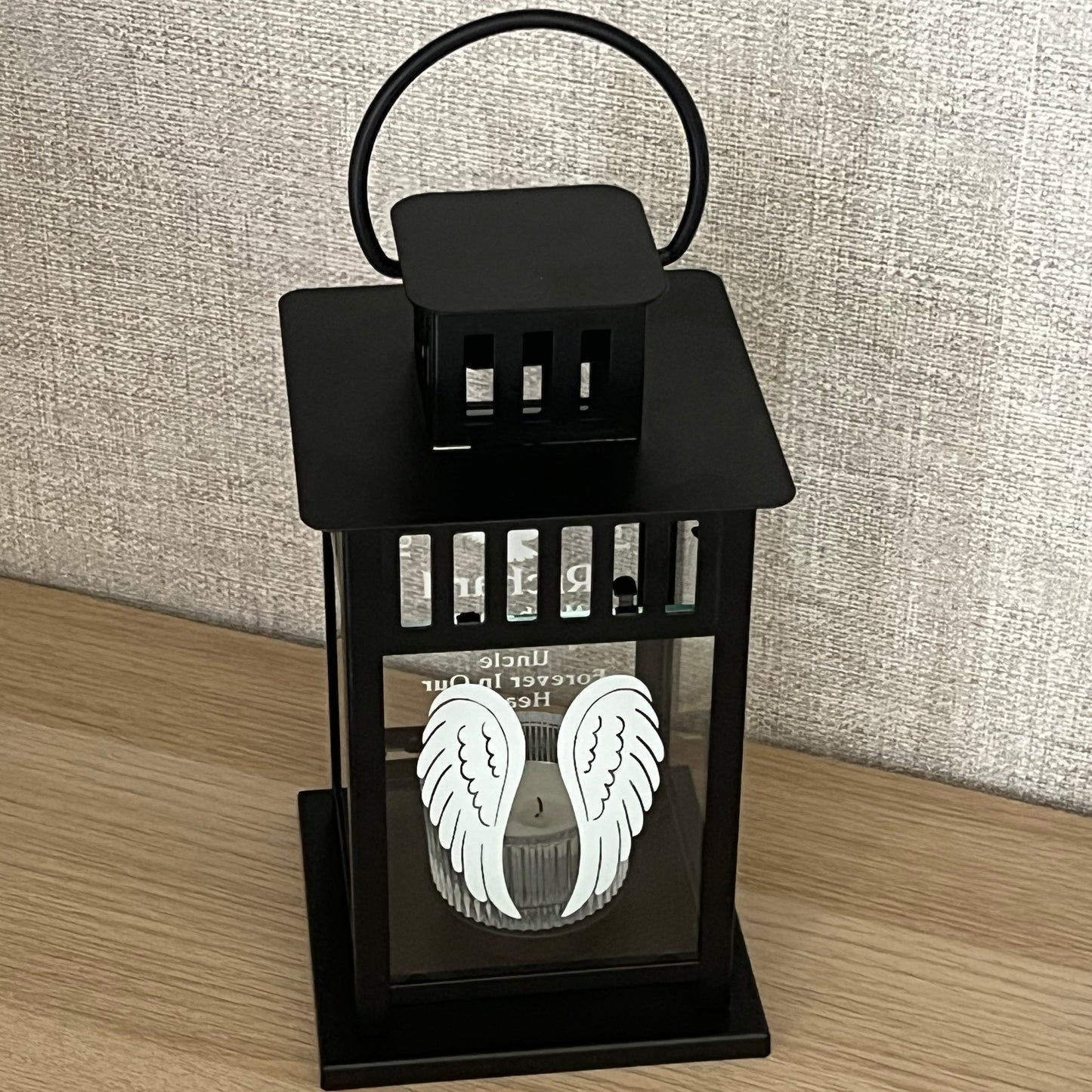 A black metal lantern with a glass window displaying a white angel wings design, resting on a light wood floor next to a textured gray rug.