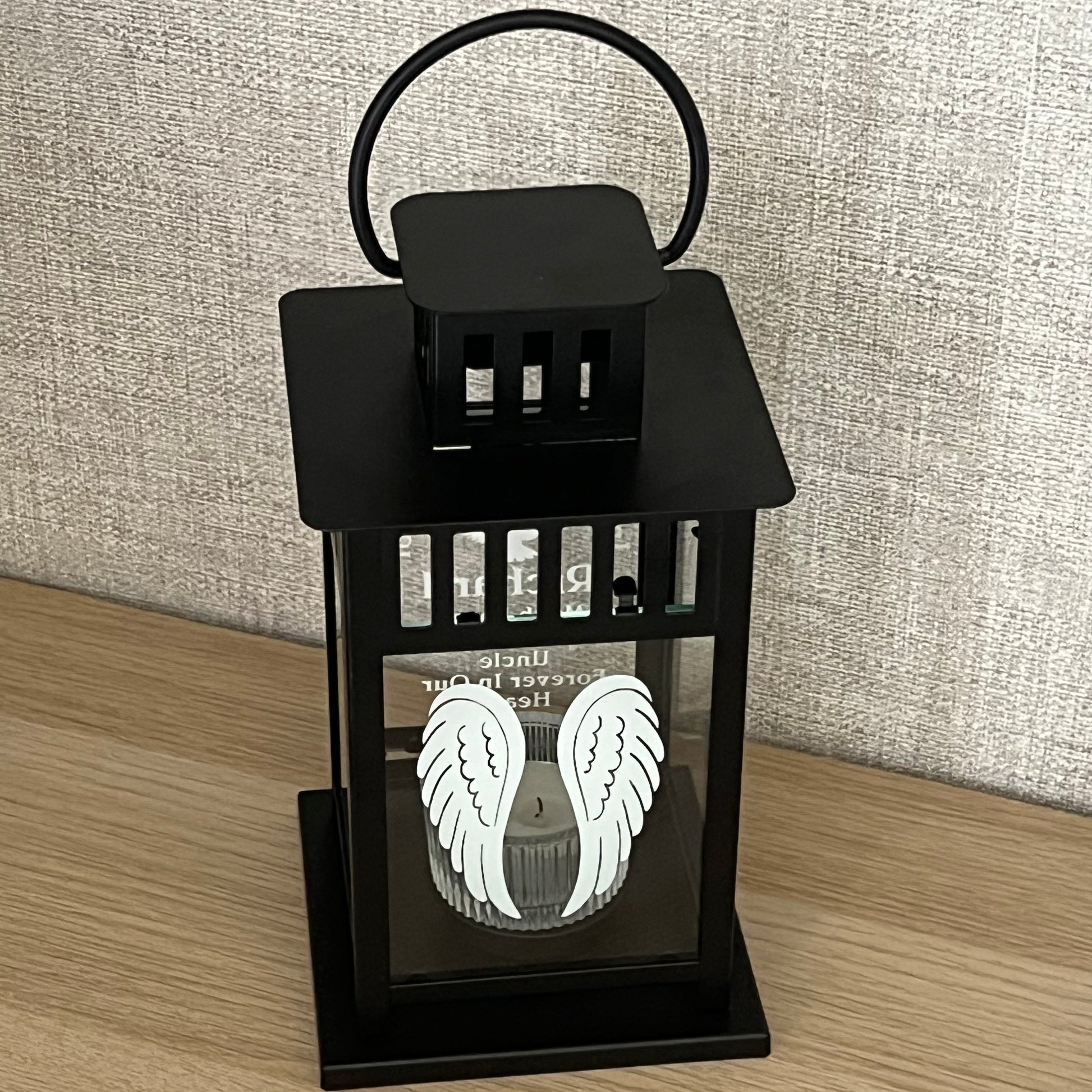 A black metal lantern with a glass window displaying a white angel wings design, resting on a light wood floor next to a textured gray rug.