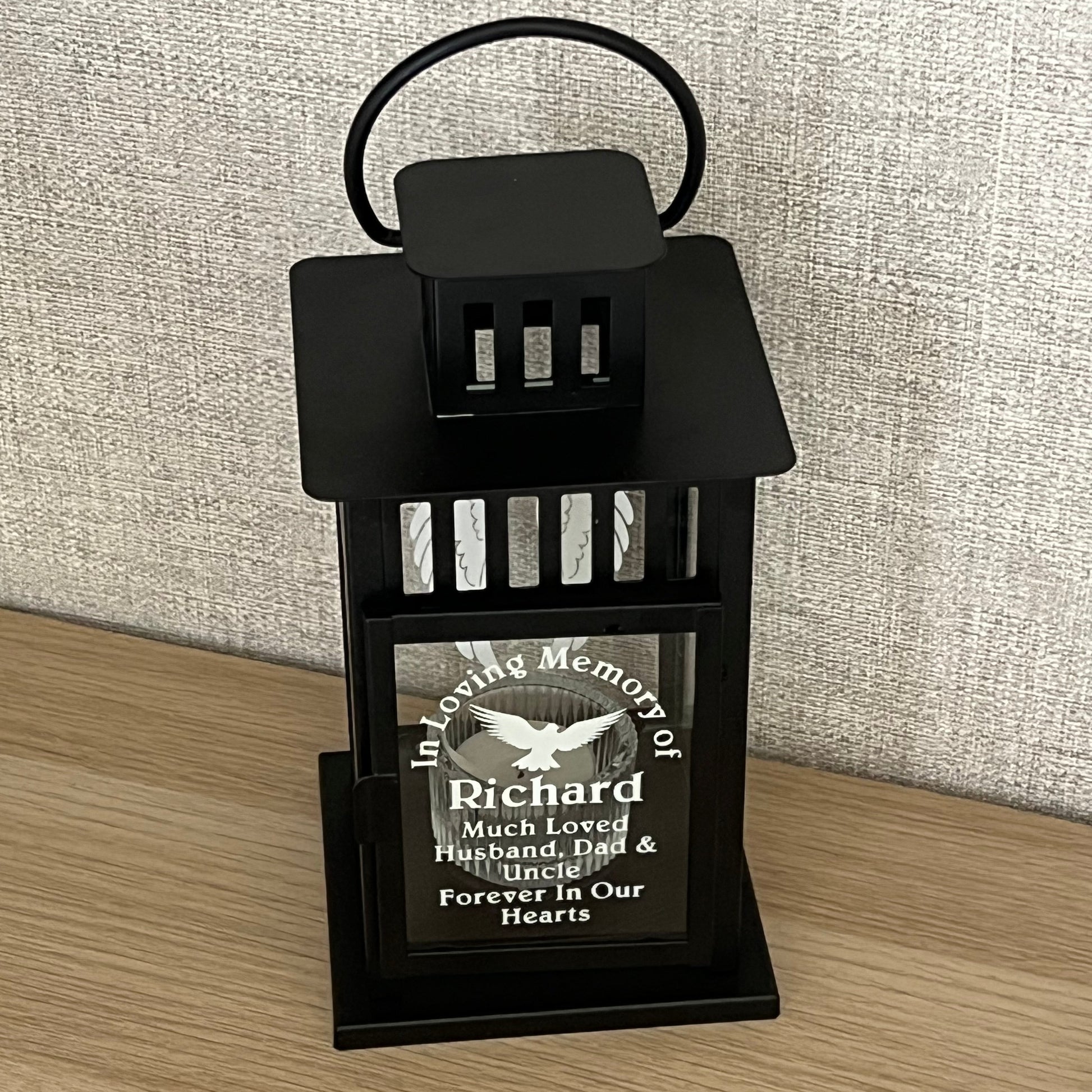 A black metal memorial urn holder with a hanging loop, shown on a light wood floor next to a textured beige fabric surface. The holder contains a small engraved black plaque with a white bird and the text 'In Memory of Richard'.
