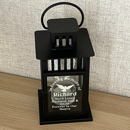 A black metal memorial urn holder with a hanging loop, shown on a light wood floor next to a textured beige fabric surface. The holder contains a small engraved black plaque with a white bird and the text 'In Memory of Richard'.