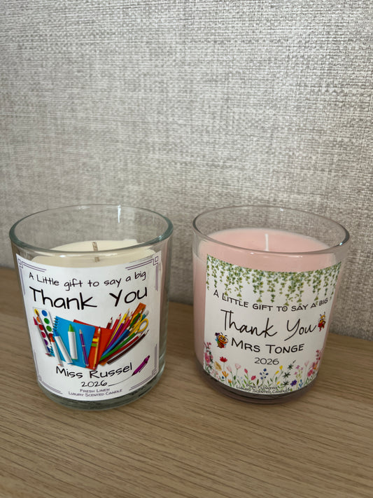 Two personalized glass jar candles on a light wood surface next to a gray textured fabric. One candle has pink wax and a floral label, and the other has cream wax with a colorful crayon and book illustration on a white label.
