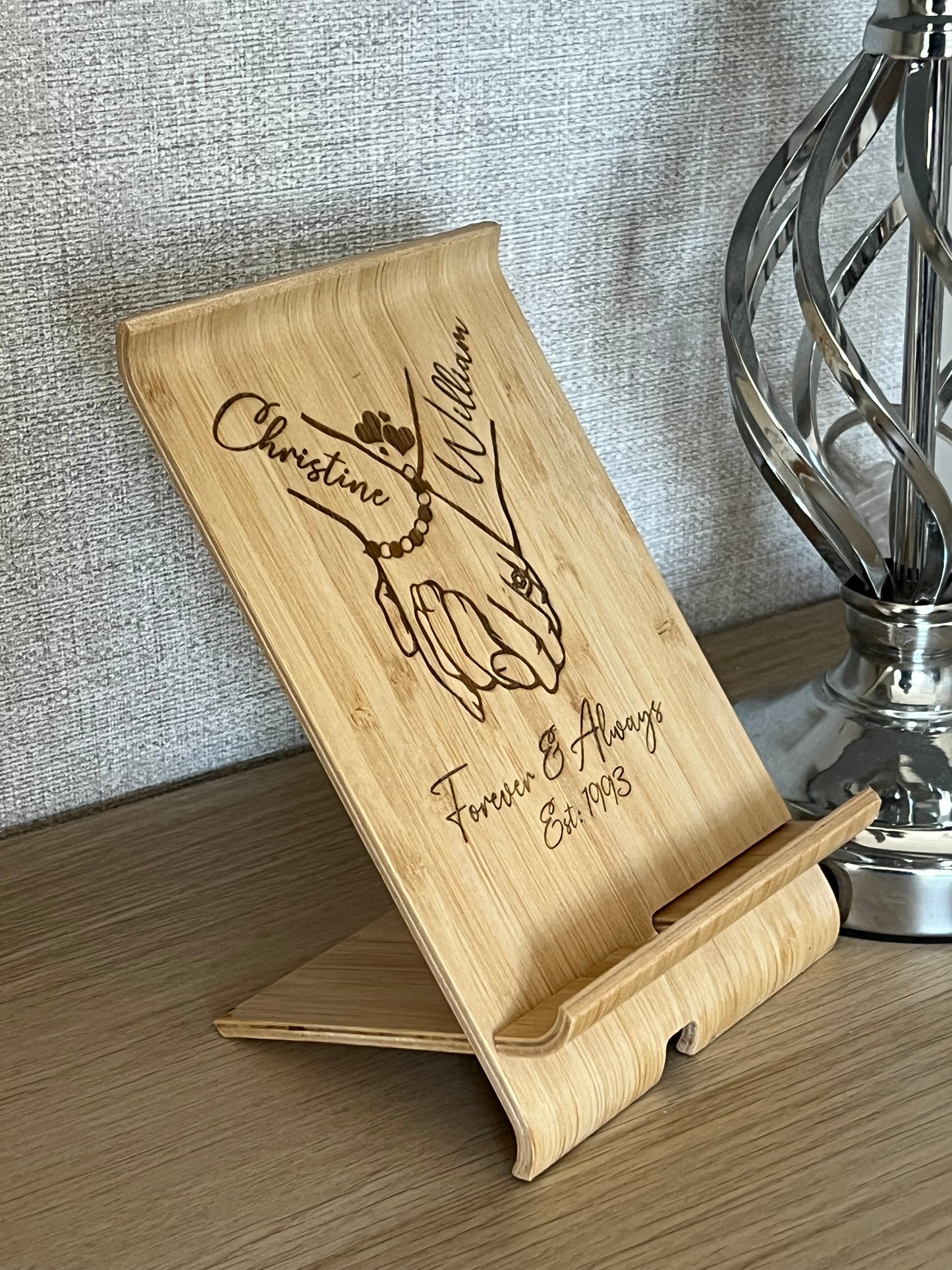A unique personalised bamboo phone stand laser-engraved with a couple holding hands, two names & a date. Perfect wedding, anniversary or Valentine's gift.