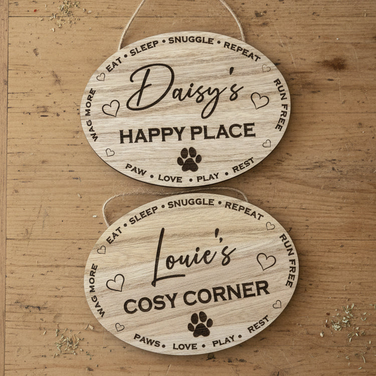  Personalised oval wooden hanging signs with  engraved text and paw prints, shown on a plain wood background. eg The top sign reads 'Daisy's Happy Place' and the bottom sign reads 'Louie's Cosy Corner'.